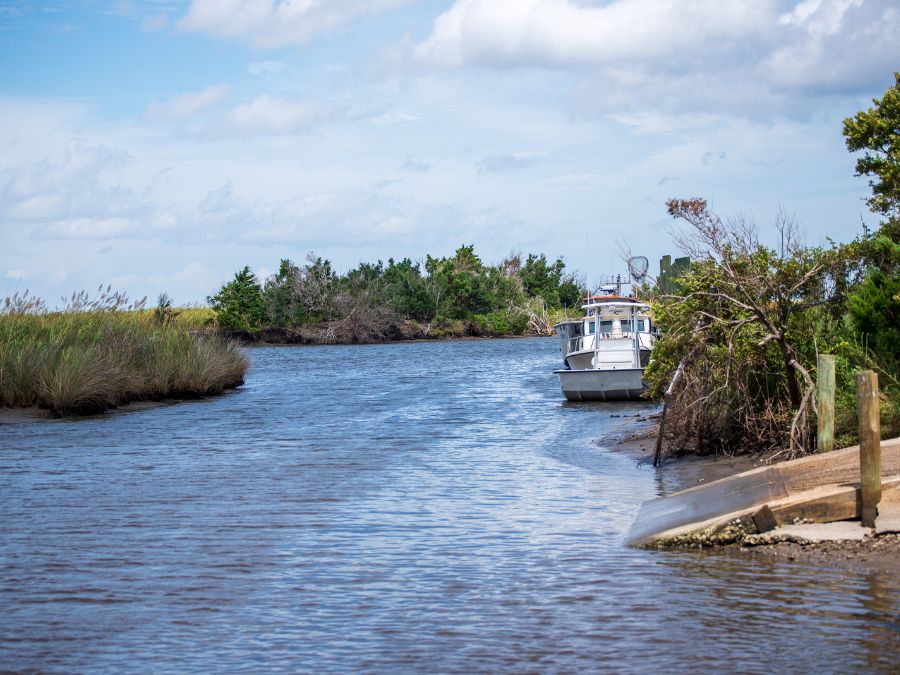 Small white boat moored along a calm coastal creek with marsh grasses, trees, and a partly cloudy sky in the background.