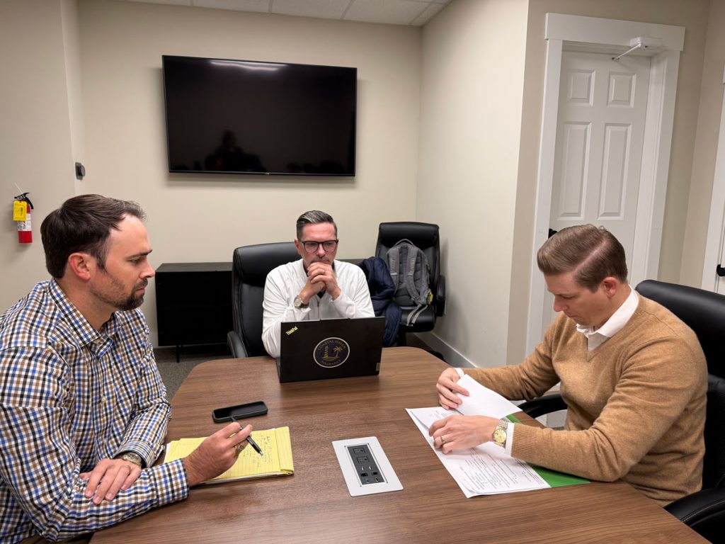 Three men in an office meeting, reviewing documents at a conference table with a laptop, notepad, and wall-mounted TV in the background.