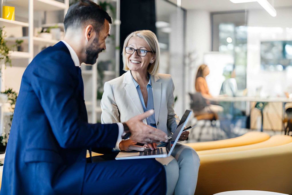 Two professionals in business attire having a friendly discussion while reviewing information on a tablet in a modern office setting.