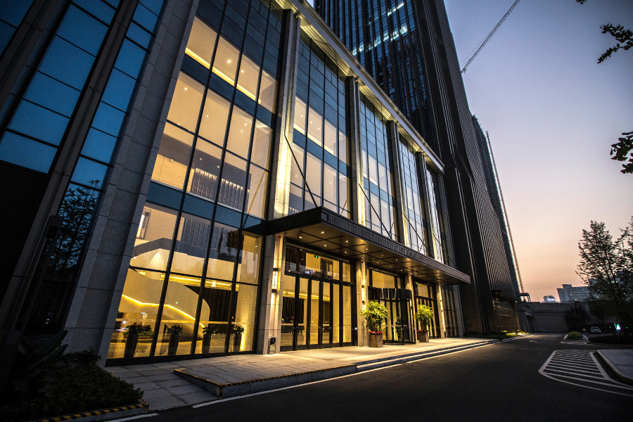 Modern glass office building entrance at dusk with illuminated lobby, tall windows, and covered drop-off area.