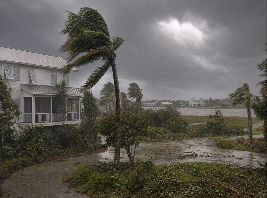 scene of a Bluffton, South Carolina neighborhood during a coastal windstorm, featuring Lowcountry-style homes with strong wind gusts bending trees and palm fronds, dark storm clouds overhead, and visible wind-driven rain.