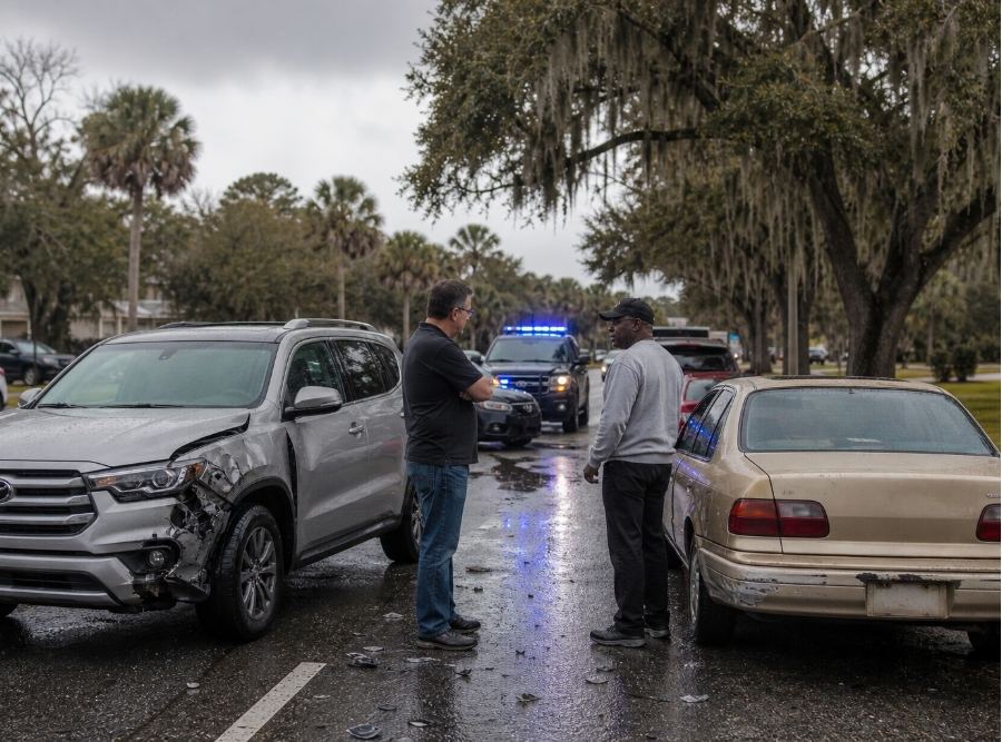 roadside scene in Bluffton, South Carolina after a multi-vehicle accident on a road resembling US-278. One vehicle shows moderate damage while another older vehicle appears uninsured. A driver stands beside their car looking concerned, speaking with another driver