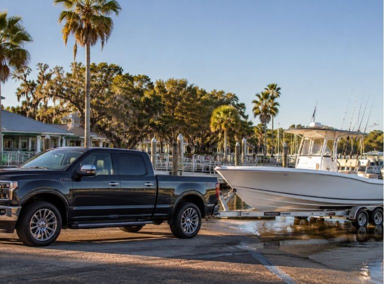 Luxury pickup truck towing a center console fishing boat at a Hilton Head Island marina with calm coastal waters, palm trees, and waterfront docks.